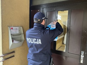 Na zdjęciu policjant wieszający plakat na drzwiach klatki schodowej.