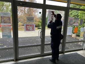 Na zdjęciu policjant przykleja na witrynie szkoły plakat.