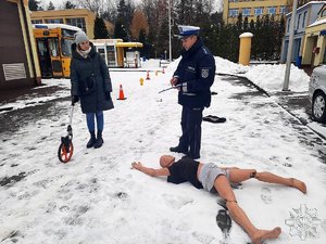 Na zdjęciu inscenizacja miejsca wypadku drogowego. policjant instruuje młoda kobietę (aplikant prokuratorski) jak prowadzić oględziny miejsca znalezienia zwłok (manekin).