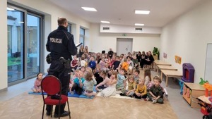 Policjant prowadzi zajęcia z dziećmi. Policjant stoi, dzieci siedzą na podłodze.