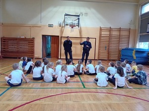 Policjanci prowadzą zajęcia z dziećmi. Dzieci zasłuchane siedzą na podłodze w sali gimnastycznej.
