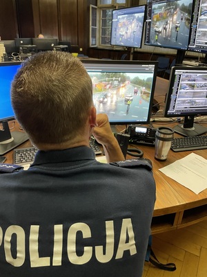 Na zdjęciu policjant siedzący przy biurku, wokół niego ekrany i monitory komputerowe na których widać policjantów zabezpieczających ulice.