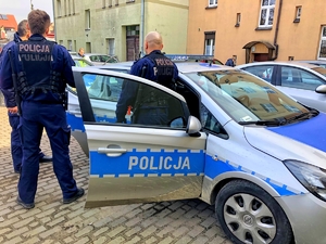 Na zdjęciu trzech policjantów stojących przy radiowozie z otwartymi drzwiami pasażera. W tle budynki kamienic.