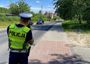 Na zdjęciu policjant mierzący miernikiem prędkość pojazdów nadjeżdżających długa prostą ulicą.