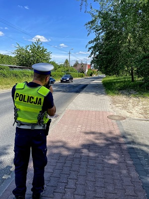 Na zdjęciu policjant mierzący miernikiem prędkość pojazdów nadjeżdżających długa prostą ulicą.