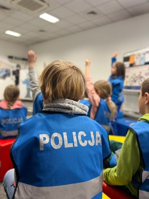 Na zdjęciu dzieci szkolne słuchające policjantki. dzieci ubrane w kamizelki z napisem policja.