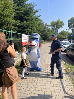 Na zdjęciu mała dziewczynka stojącą naprzeciwko maskotki policyjnej Psa Sznupka, obok policjant w mundurze.