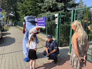 Na zdjęciu mała dziewczynka stojącą naprzeciwko maskotki policyjnej Psa Sznupka, obok kucający i rozmawiający z nią policjant w mundurze.