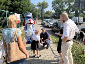 Na zdjęciu mała dziewczynka stojącą naprzeciwko maskotki policyjnej Psa Sznupka, obok kucający policjant w mundurze. Rozmowie przysłuchują się rodzice odprowadzający dziecko do szkoły.