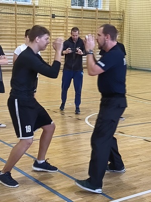 Na zdjęciu policjant i uczeń naprzeciwko siebie w pozach obronnych z uniesionymi i zasłaniającymi twarze ramionami.