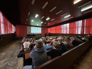 Na zdjęciu aula wypełniona słuchaczami. Na scenie wyświetlany jest slajd z napisem Debata. Widać siedzących tam prelegentów.