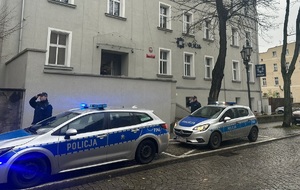 Na zdjęciu dwa radiowozy i dwóch policjantów w pozycji wyprostowanej salutując.