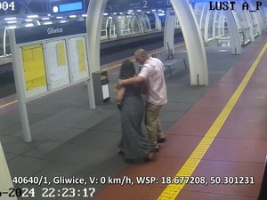 Na zdjęciu mężczyzna  w różowej koszuli i jasno niebieskich spodniach, stojący na peronie dworca wraz z kobietą w długich włosach stojącą tyłem.