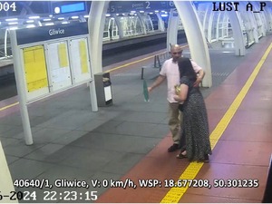 Na zdjęciu mężczyzna  w różowej koszuli i jasno niebieskich spodniach, stojący na peronie dworca wraz z kobietą w długich włosach stojącą tyłem.