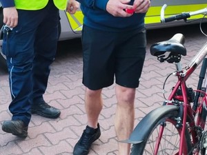 stojący rower, rowerzysta oraz policjant umundurowany (dolne części ciała). W tle widać radiowóz