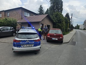 Radiowóz oznakowany i policjant umundurowany z zatrzymaną. W tle znajdują się budynki.