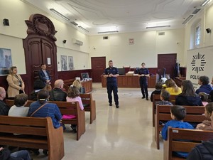 Zajęcia na sali sądowej prowadzi umundurowany policjant. W ławkach siedzą zwiedzającvy