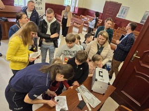 Grupa zwiedzających stoi na sali sądowej. Policjantka umundurowana prezentuje zbuieranie odcisków palców