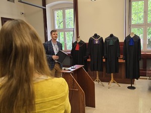 Na sali sądowej stoi osoba prowadząca wykła. Na środku pokoju stoi ekspozycja: trzy różne togi.