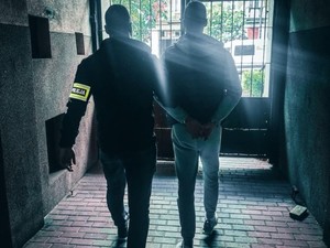 Nieumundurowany policjant z opaską na ręku prowadzi w bramie zatrzymanego