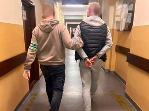 Nieumundurowany policjant w opasce na ręku prowadzi zatrzymanego korytarzem