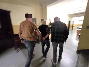 Dwóch nieumundurowanych policjantów z opaskami POLICJA prowadzi korytarzem zatrzymanego