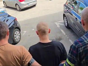 Dwóch nieumundurowanych policjantów z opaskami POLICJA prowadzi zatrzymanego. Wychodzą z budynku.
