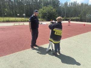 policjant umundurowany stoi z chłopcem ubranym w kurtkę Straży Pożarnej