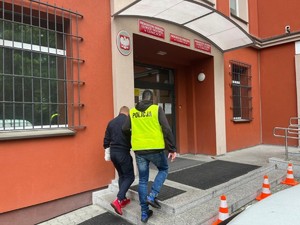 Policjant w kamizelce prowadzi zatrzymanego. Wchodzą do budynku Prokuratury.
