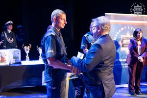 Na zdjęciu policjant odbierający nagrodę na scenie w konkursie od mężczyzny w cywilnym ubraniu.