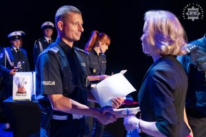 Na zdjęciu policjant odbierający nagrodę na scenie w konkursie od kobiety w cywilnym ubraniu.