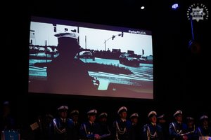 Przyciemniona sala, na planszy wyświetlony film. Na scenie stoi orkiestra policyjna.