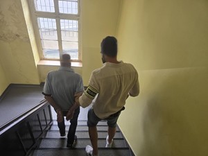 Na zdjęciu nieumundurowany policjant z opaską z napisem Policja prowadzi zatrzymanego po schodach