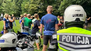 Na zdjęciu widać plecy policjanta siedzącego na motocyklu w kamizelce odblaskowej. Obok stoi grupa ludzi.