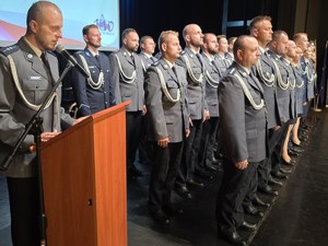 Na scenie umundurowany policjant przy mównicy, policjanci gotowi do wręczenia aktów mianowania.