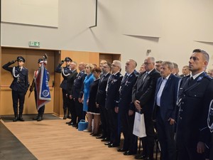 Poczet sztandarowy stoi pod ścianą. Ludzie na dużej sali stoją.