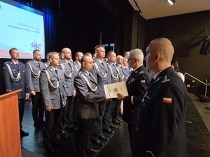Na scenie umundurowany policjant przy mównicy, policjanci gotowi do wręczenia aktów mianowania.