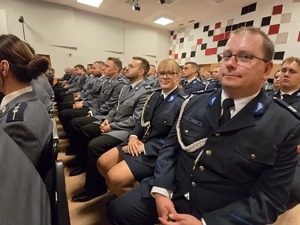 Ludzie siedzący na krzesłach na dużej sali, zwróceni w kierunku sceny.