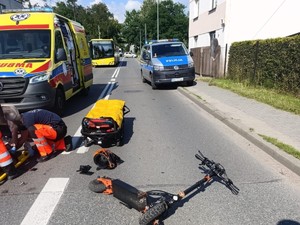 Na zdjęciu radiowóz Policji i Pogotowia ratunkowego, obok ratowników, którzy udzielają pomocy stoją nosze. Na jezdni leży hulajnoga.