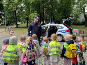 Grupa dzieci w wieku przedszkolnym stoi przy otwartym radiowozie. Policjant umundurowany prowadzi pogadankę z dziećmi.