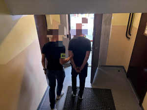 Nieumundurowany policjant z opaską na ręku prowadzi zatrzymanego w kajdankach.. Wychodzą z budynku.