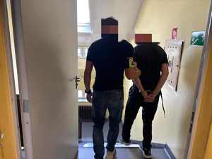 Nieumundurowany policjant z opaską na ręku prowadzi zatrzymanego w kajdankach. Schodzą po schodach.