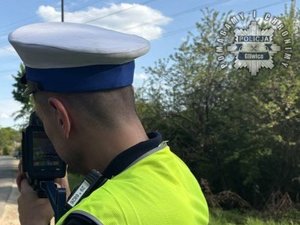 Na zdjęciu policjant ruchu drogowego w białej czapce i kamizelce odblaskowej podczas pomiaru prędkości pojazdów.