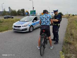 Kontrola rowerzysty przez policjanta ruchu drogowego. W tle stoi radiowóz oznakowany.