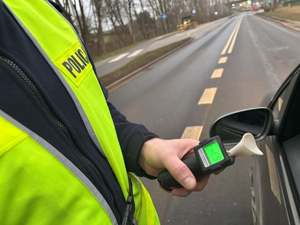 Zdjęcie policjanta w żółtej kamizelce odblaskowej z urządzeniem do badania stanu trzeźwości. Policjant stoi na jezdni przy pojeździe.