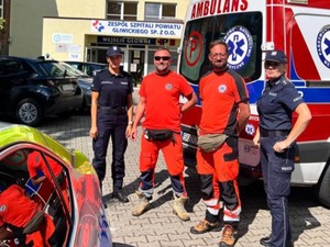 Na tle karetki pogotowia stoi dwóch ratowników i dwie policjantki. Z boku fragment radiowozu.