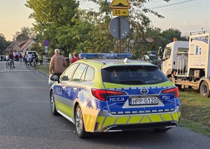 Radiowóz oznakowany Policji jadący ulicą. W tle widać osoby spacerujące i zaparkowane pojazdy.