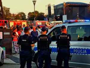Trzech policjantów stoi przy radiowozie oznakowanym. W tle scena i tłum ludzi.