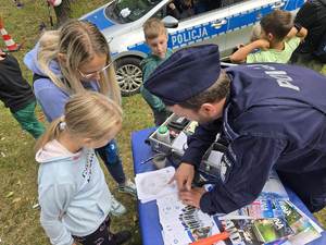 Policjant przy stoisku profilaktycznym pokazuje dzieciom pobieranie linii papilarnych dłoni.