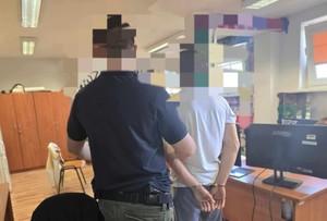 Na zdjęciu wewnątrz pomieszczenia widać od tyłu policjanta nieumundurowanego trzymającego za ramię zatrzymanego, który jest w kajdankach.